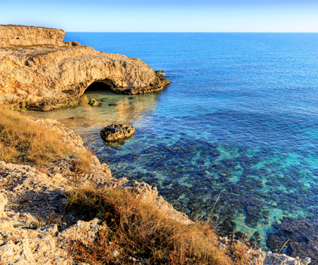 Zee kabbelt tegen rotsige kustlijn bij Paralimni, Cyprus, onder heldere hemel. Blauwe en turquoise tinten domineren.